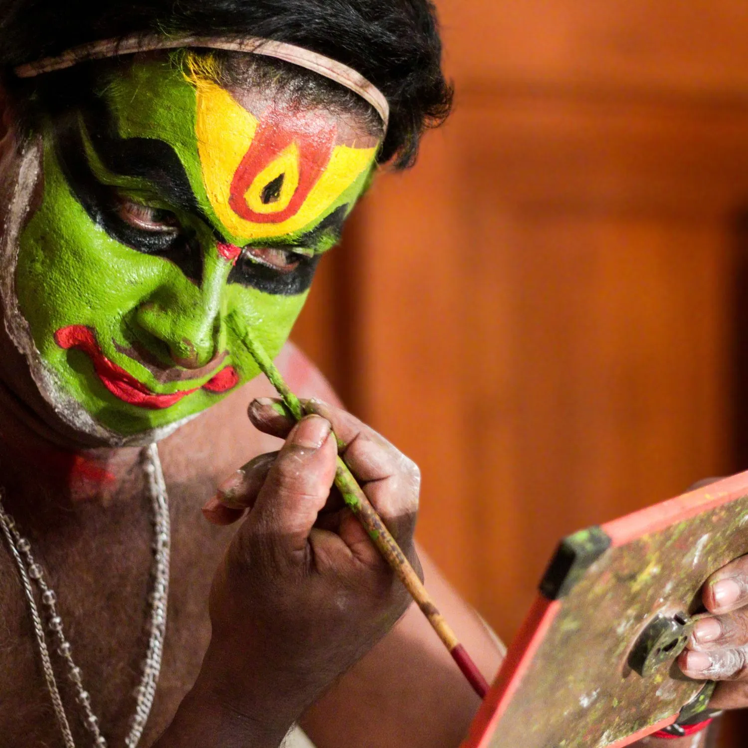Kathakali Artist Preparing for a Traditional Performance in Retreat Inn Kerala Ayurveda Yoga Resort. A traditional Kathakali artist meticulously applying vibrant green and red face paint while looking into a mirror, preparing for a classical performance in Retreat Inn Kerala Ayurveda Yoga Resort, Kerala, India.