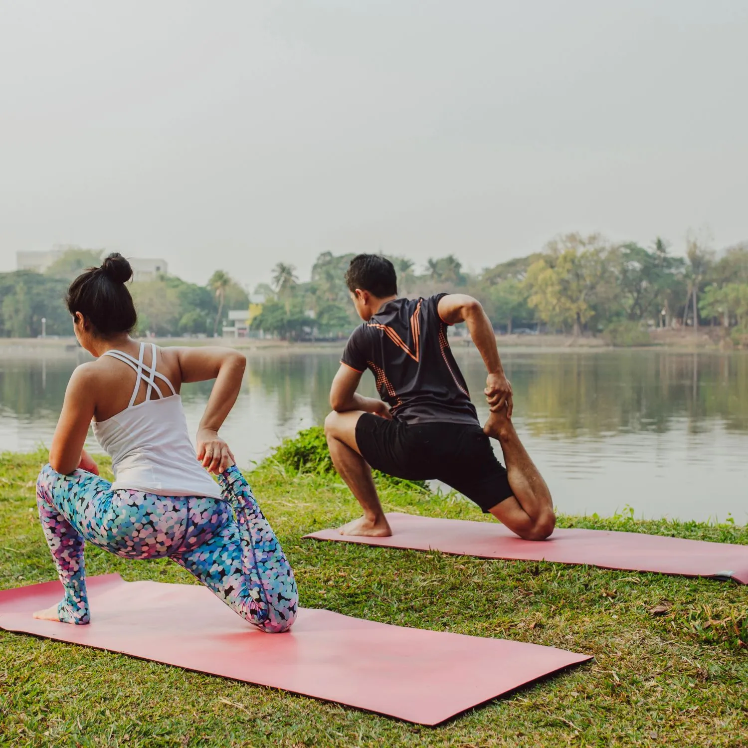 couple-doing-yoga-with-beautiful-landscape- Retreat Inn Kerala Ayurveda Yoga Resort. A man and a woman practicing yoga on red mats near a serene lakeside, surrounded by lush greenery and distant hills, under a clear sky at Retreat Inn Kerala Ayurveda Yoga Resort.