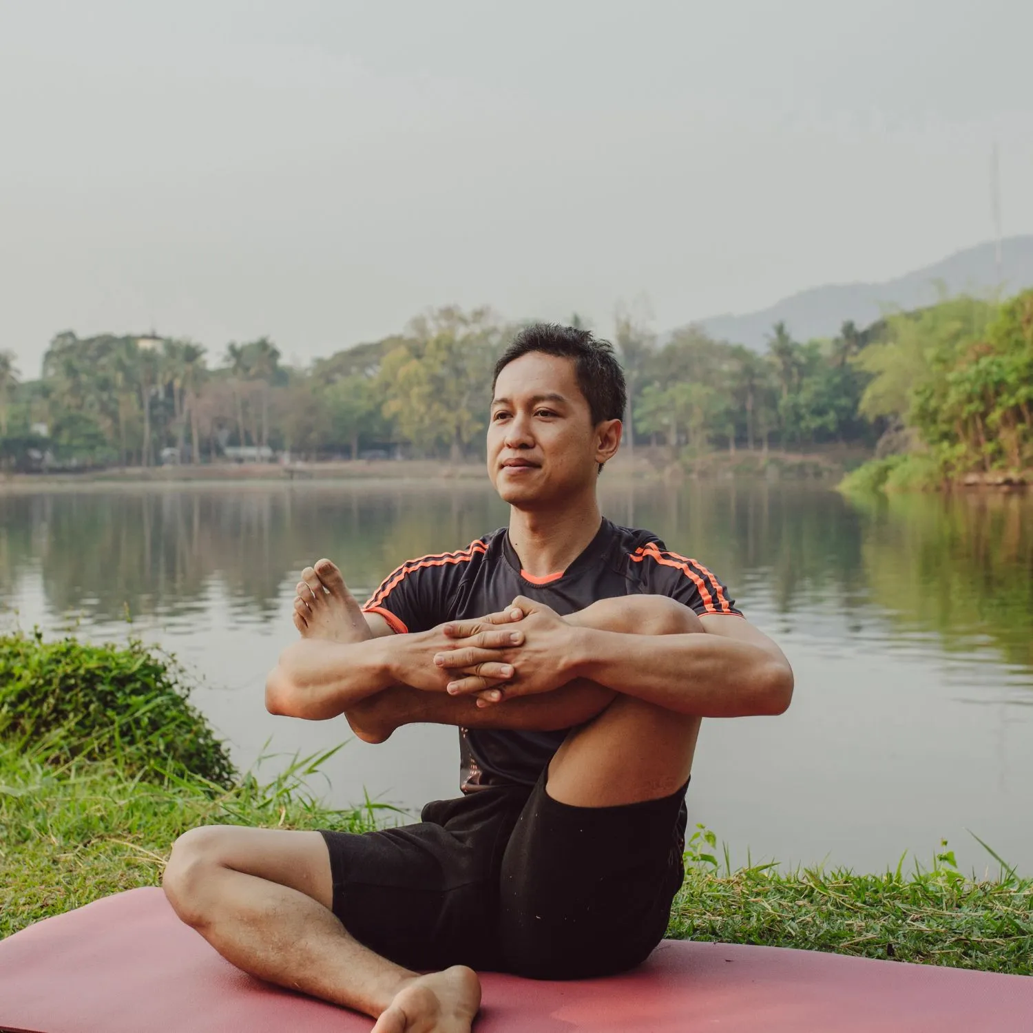 periyar river side yoga at retreat inn kerala ayurveda yoga resort Man practicing yoga by the lakeside, performing a seated asana on a red mat amidst serene natural surroundings at retreat inn Kerala ayurveda yoga resort.