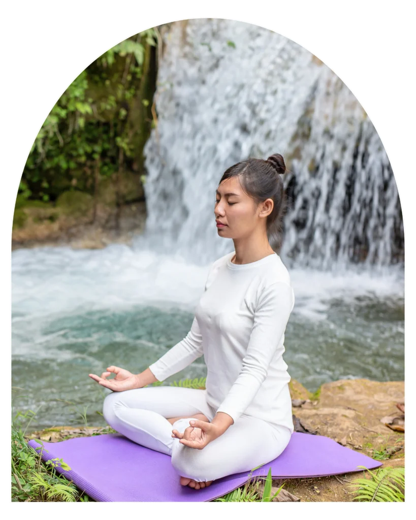 Person meditating in lotus pose on a yoga mat near a natural waterfall in a peaceful forest setting during a wellness treatment in Retreat Inn Kerala Ayurveda Yoga Resort